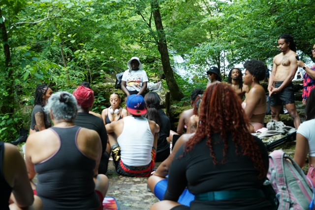 Group of people sitting and standing in a forested area with green trees, some wearing swimsuits or casual clothing.