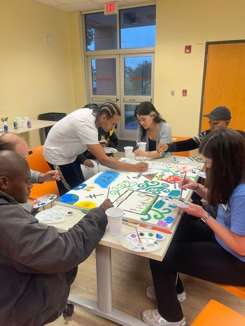 People gathered around a table painting colorful designs on paper, in a room with yellow walls and large windows.