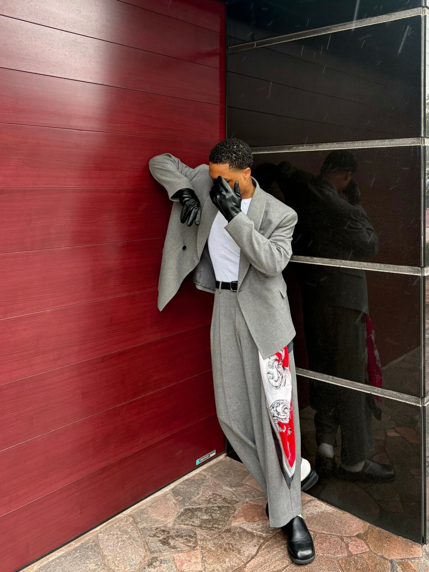 Person in gray coat and patterned pants covers face with hand, leaning against red wall, with black and reflective surfaces nearby.