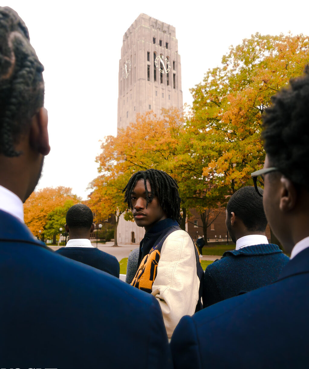 Group of students in blue uniforms outdoors with a historic tower and autumn trees in background.