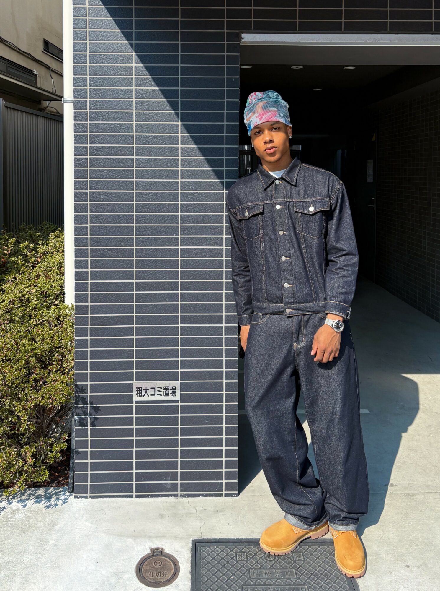 Young person standing outdoors next to a tiled wall, wearing a denim jacket, baggy jeans, colorful headscarf, and tan shoes.