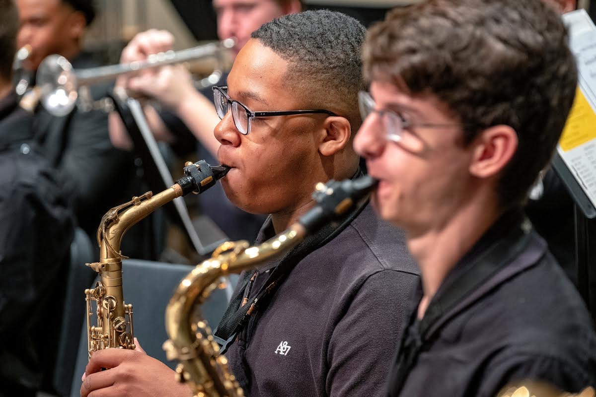 Two musicians playing saxophones in an orchestra, focusing on their faces and instruments, with other musicians in the background.