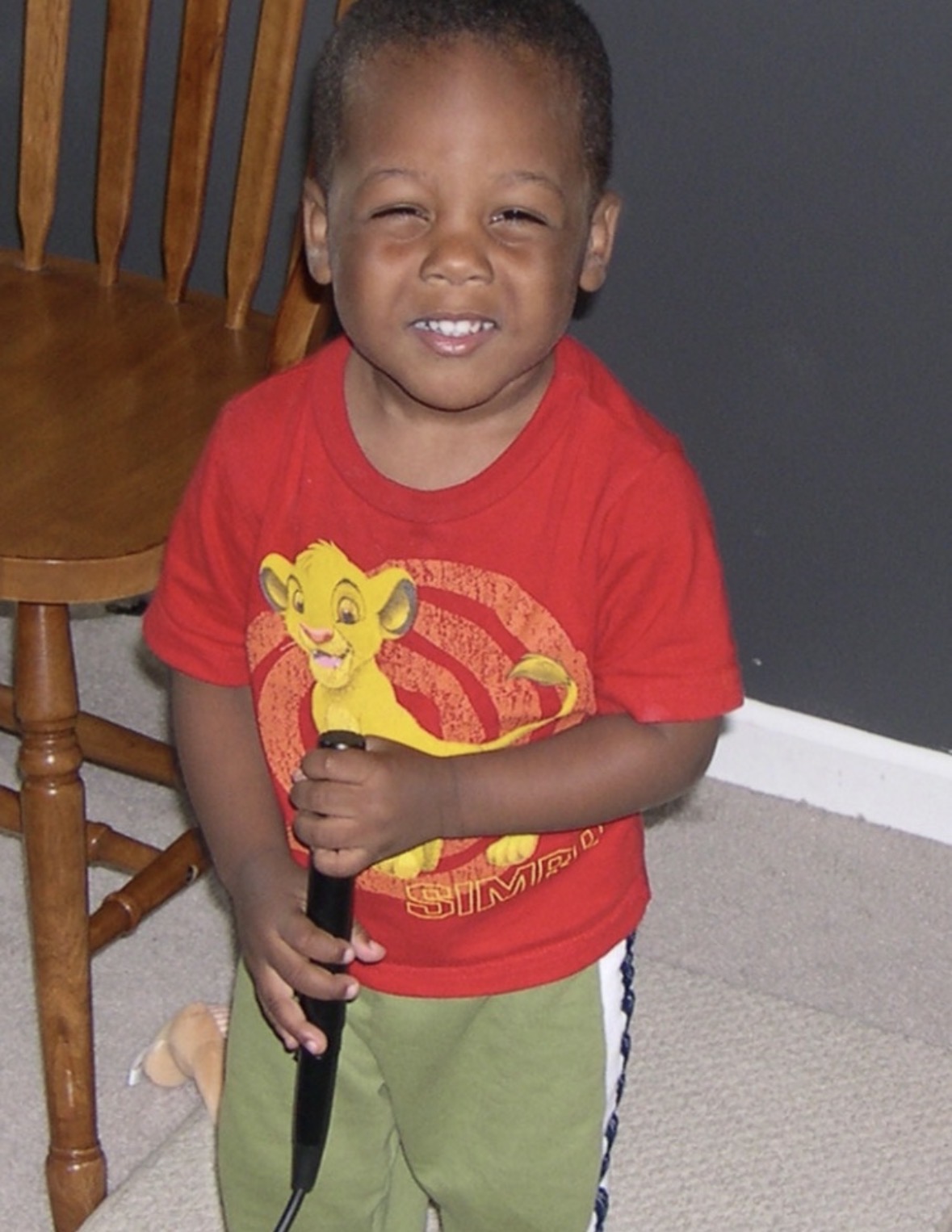 Smiling young child in red shirt holding a microphone, standing indoors near a wooden chair and gray wall.