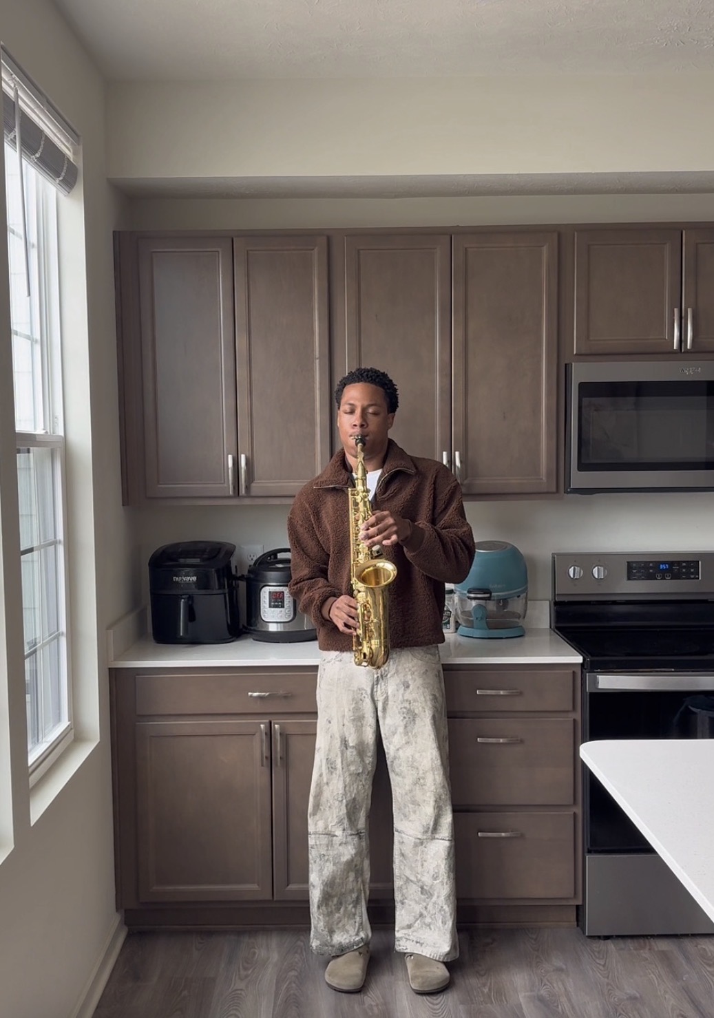 Person standing in kitchen playing saxophone, cabinets, microwave, and appliances visible, natural light from window.