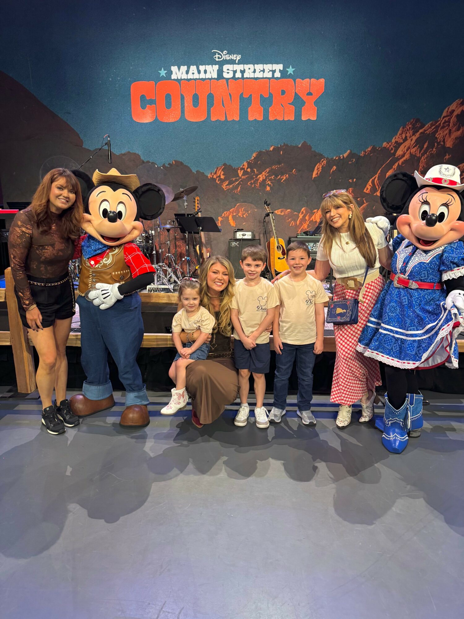 Group of children and adults with Mickey Mouse and Minnie Mouse characters at a themed event, backdrop shows mountains and 'MANY STREET COUNTRY' text.
