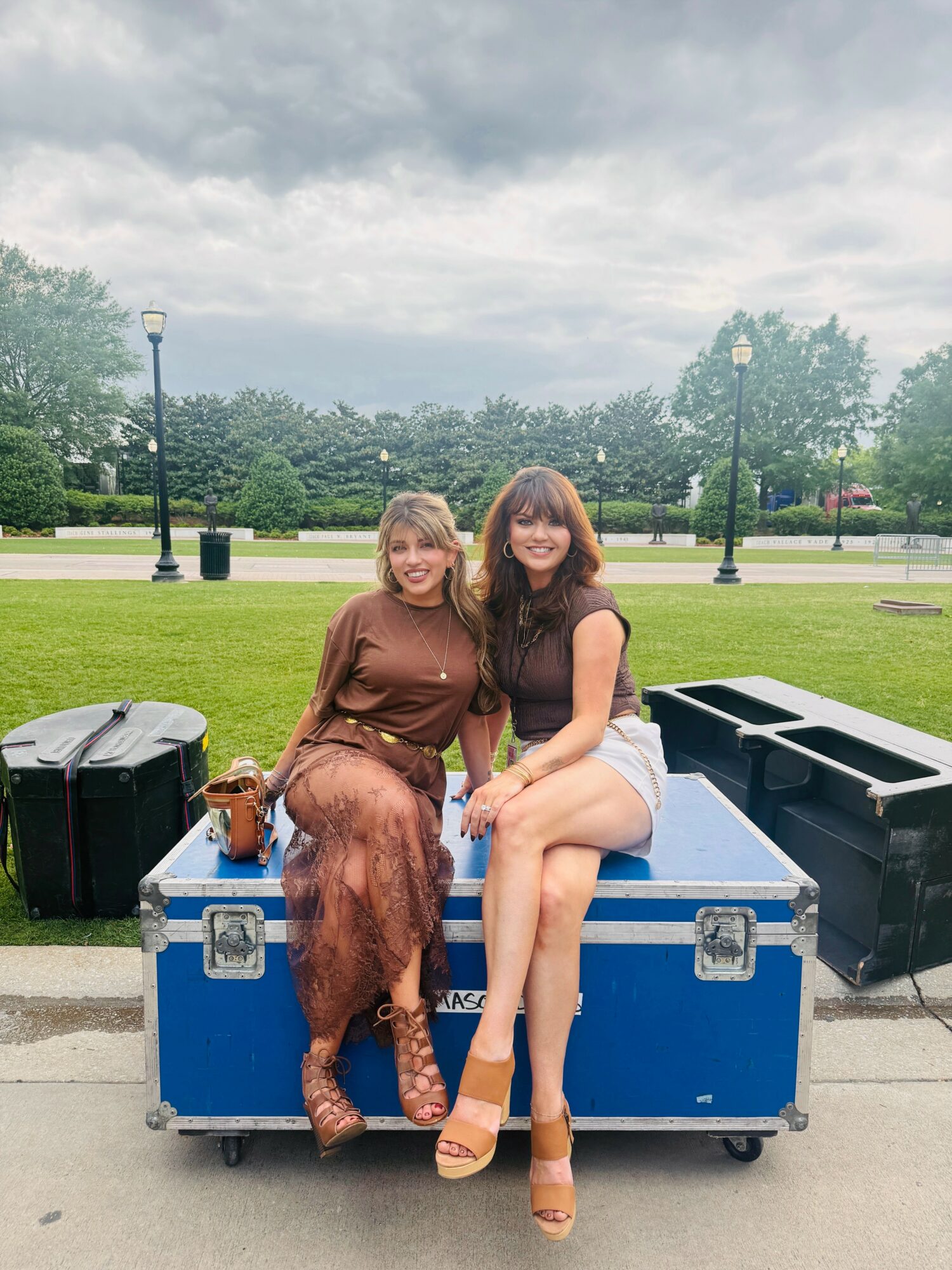 Two women sitting on a blue equipment case outdoors, smiling, with a park and cloudy sky in background.