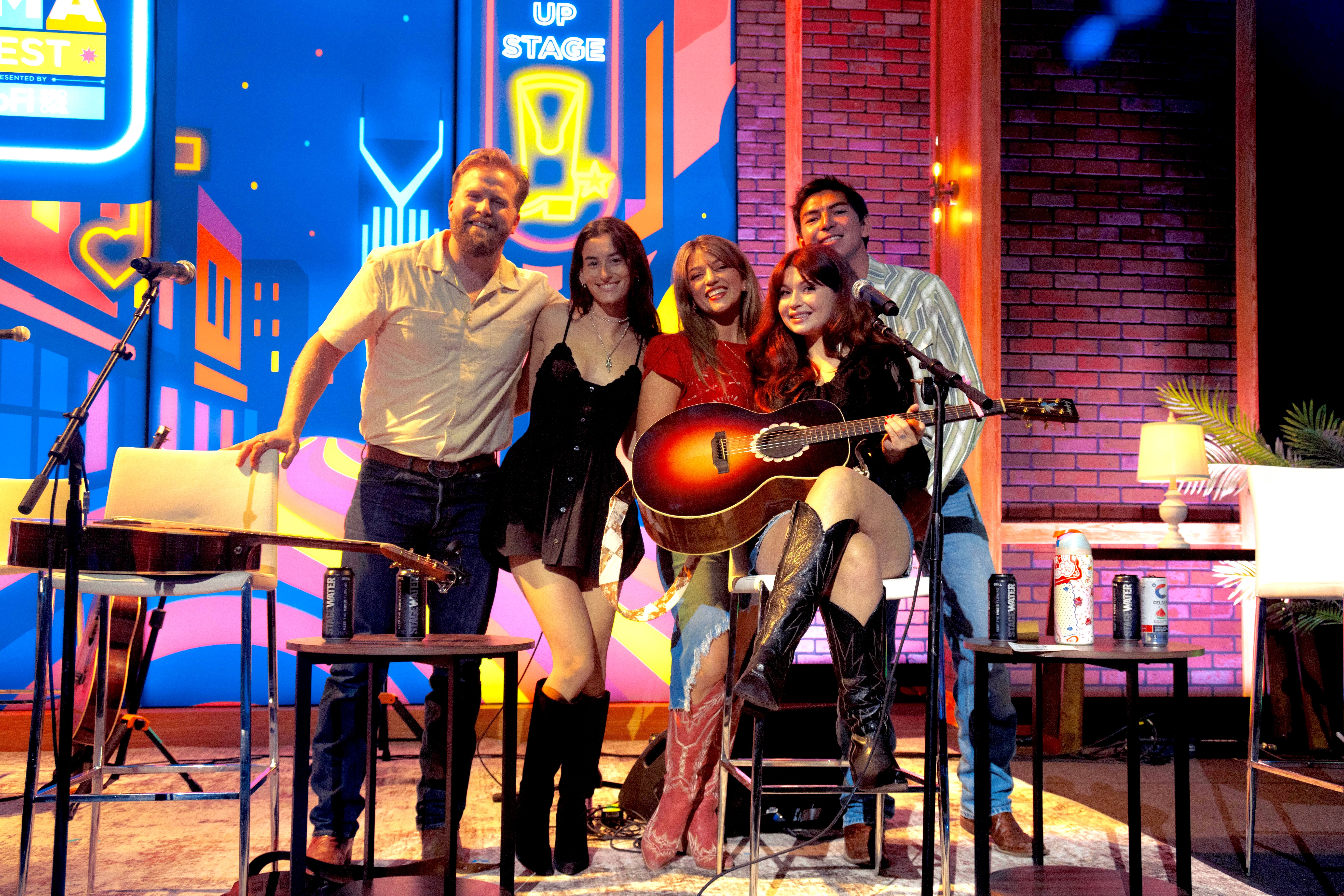 Group of five people on a colorful stage, one holding a guitar, smiling, with musical equipment and cityscape background.