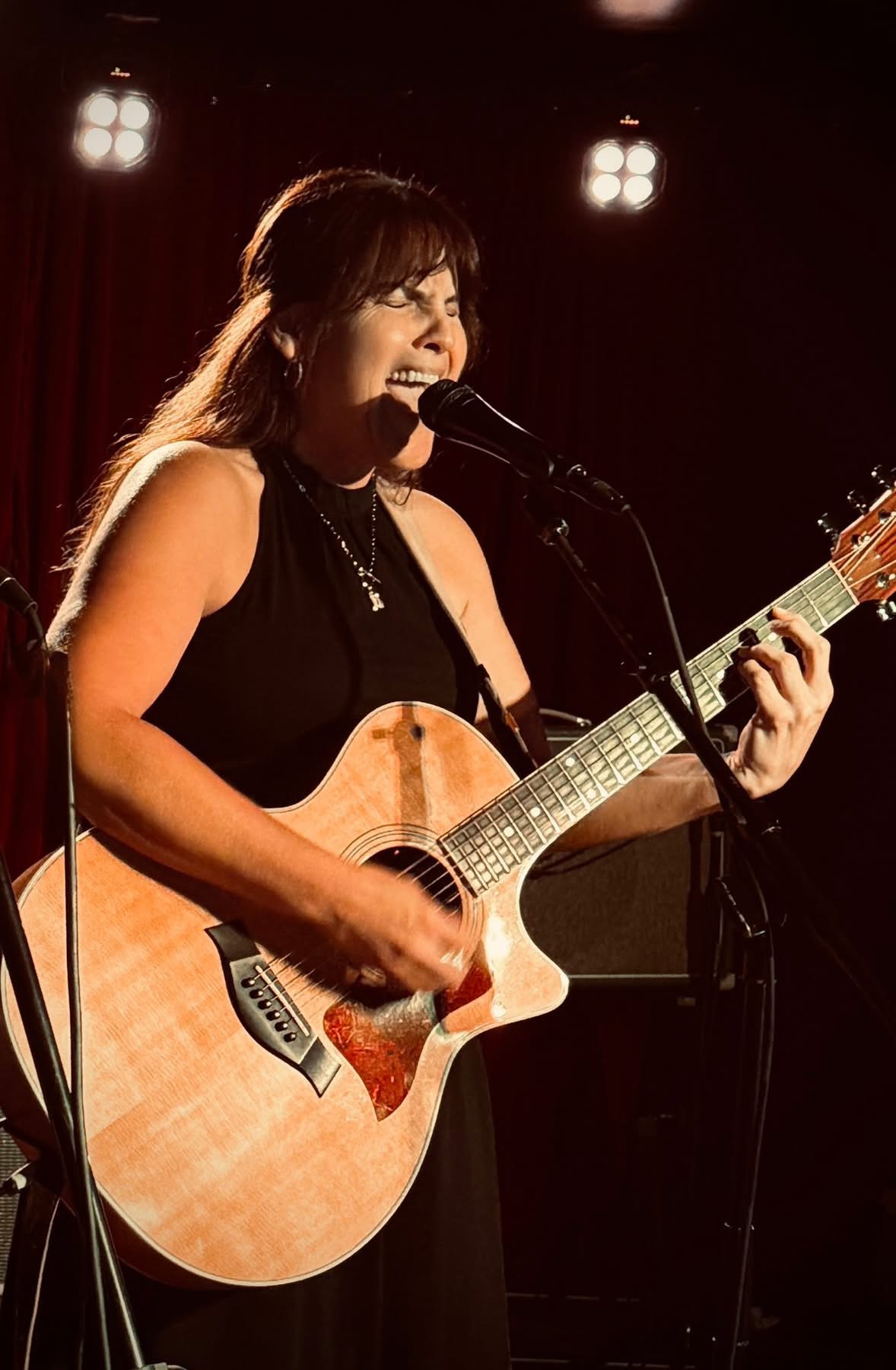 Woman singing and playing guitar on stage with bright lights behind her.