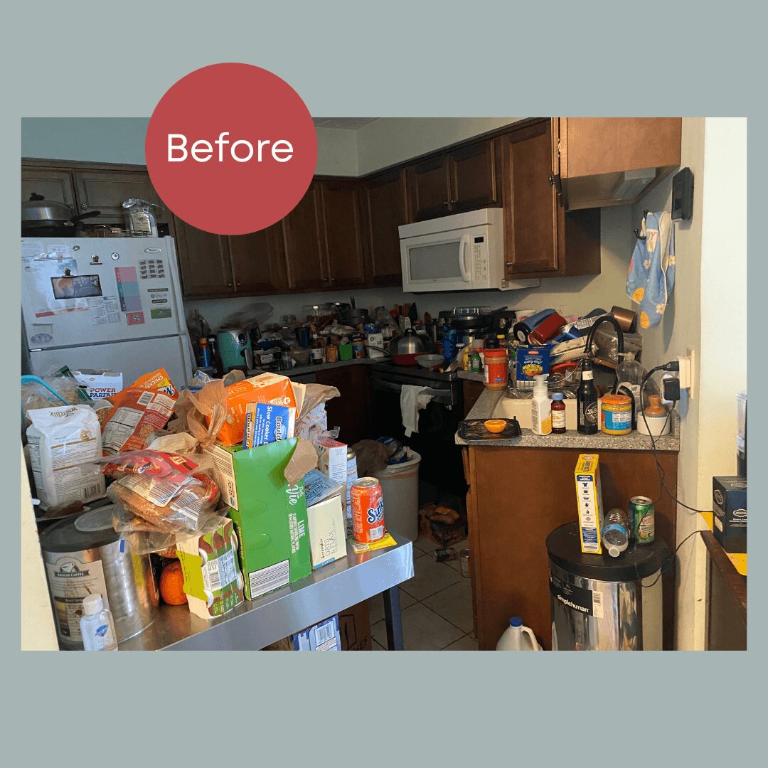 Cluttered kitchen with food, bottles, and appliances on counters and table, microwave above stove, and cabinets in background.