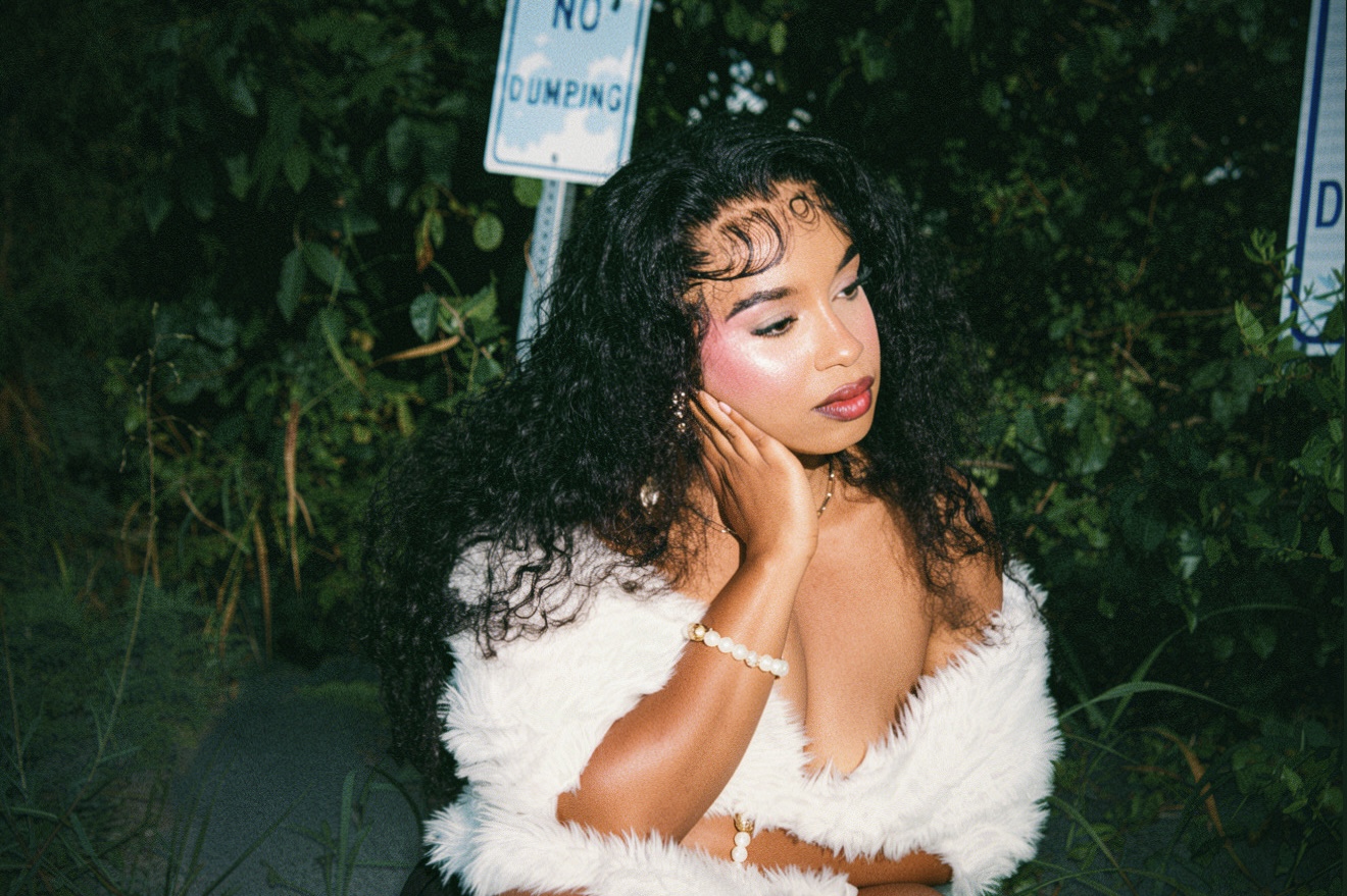 Young woman with curly hair and makeup, wearing a white fur coat, outdoors at night with greenery and signs behind her.