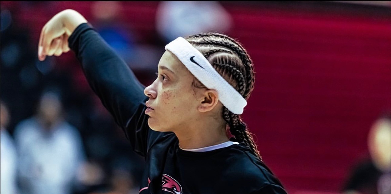 Female athlete with braided hair and a white headband, preparing to serve in a sports event, with a blurred background.