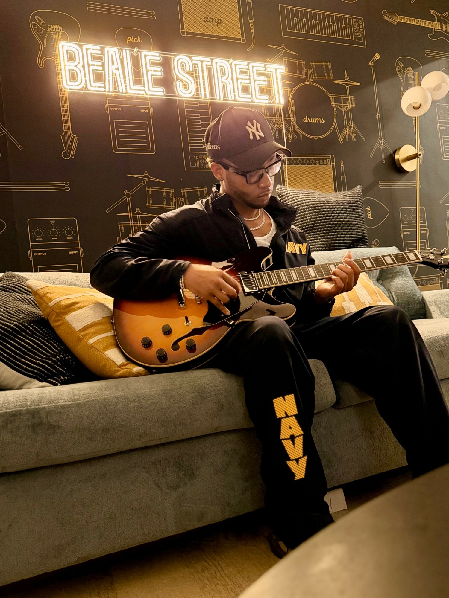 Person sitting on a sofa playing an electric guitar, wearing a cap and glasses, with a neon sign and wall art behind.