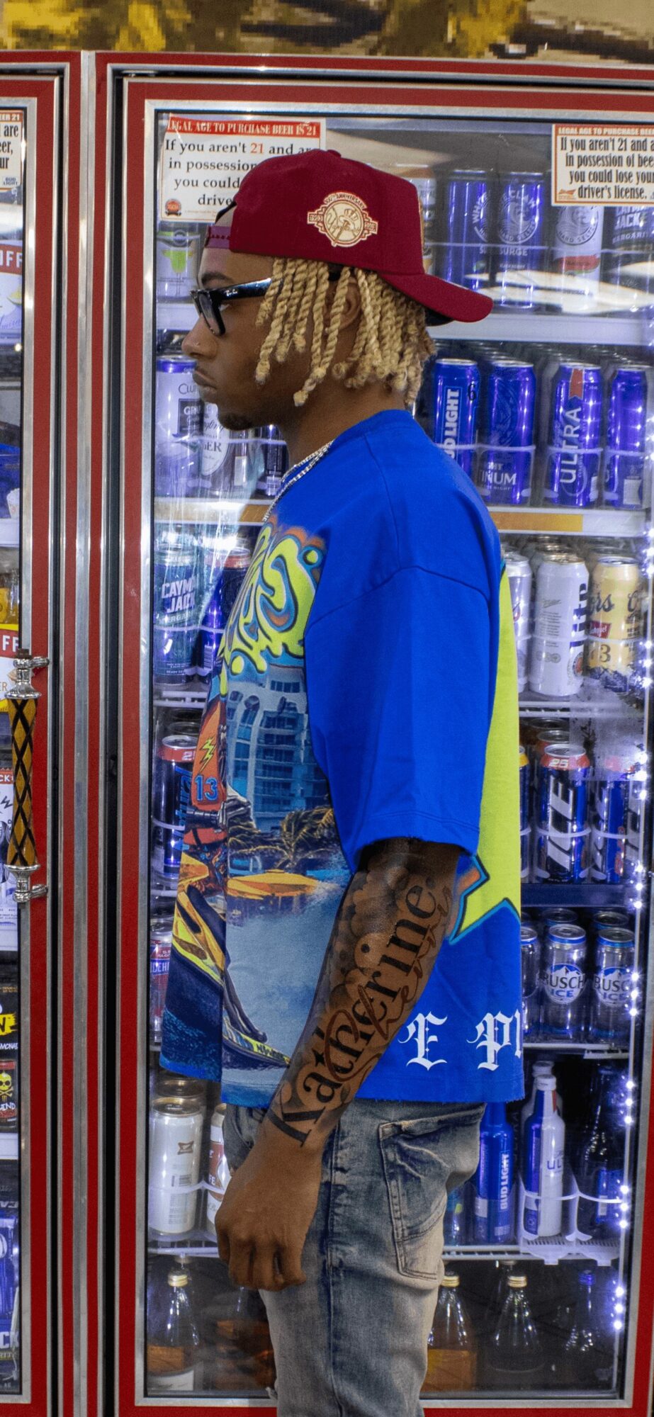 Young man with blonde dreadlocks wearing a red cap, sunglasses, and a blue shirt, standing in front of a vending machine.