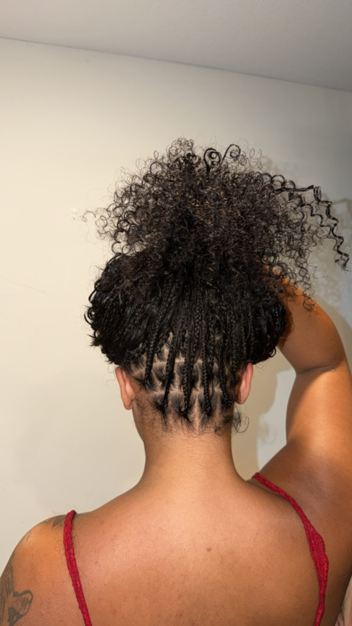 Back view of a person with curly hair styled in a high puff, wearing a red strap top, against a plain wall.