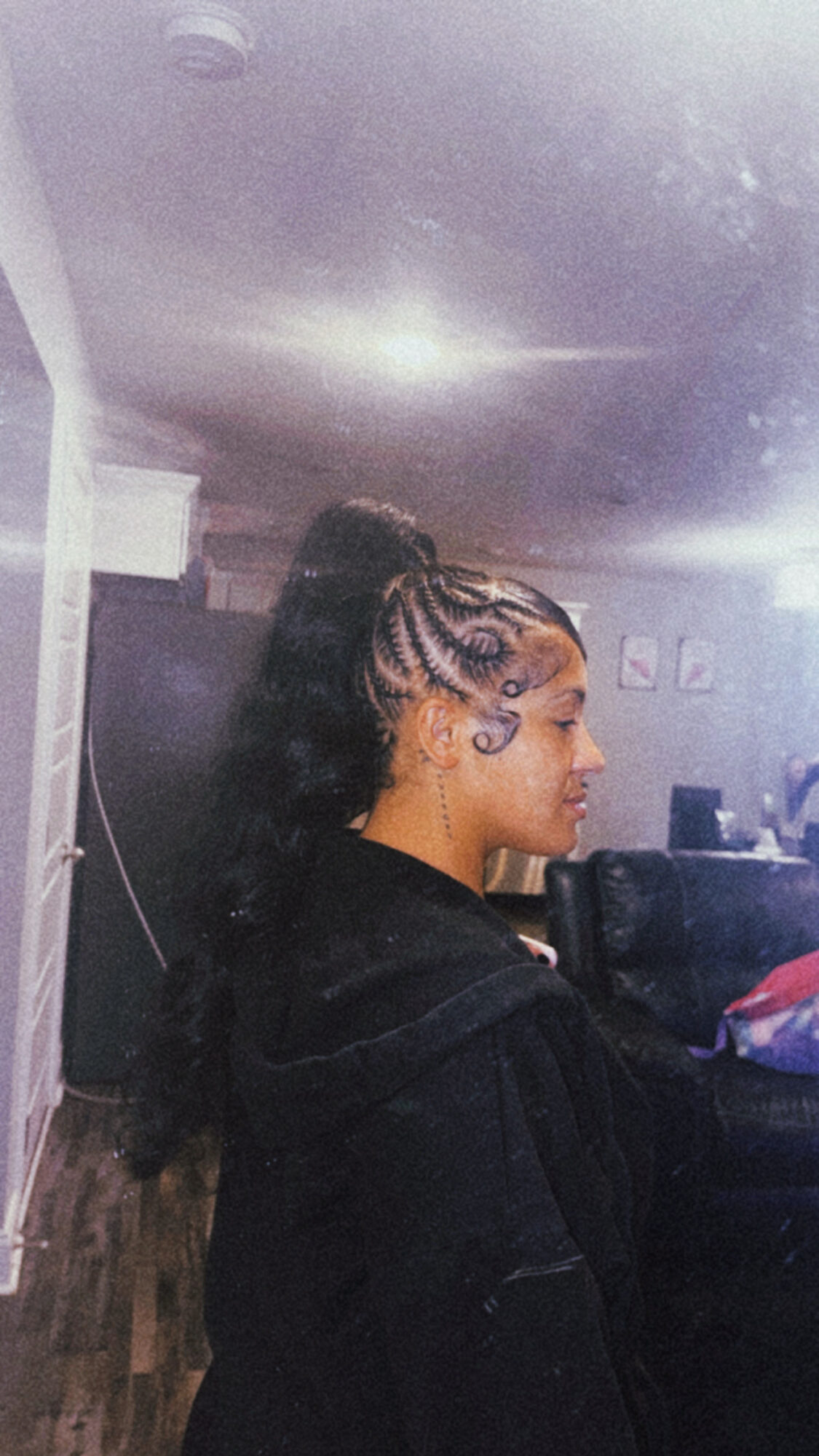 Side view of a woman with braided hairstyle in a room, taking a mirror selfie, with a dark hoodie.