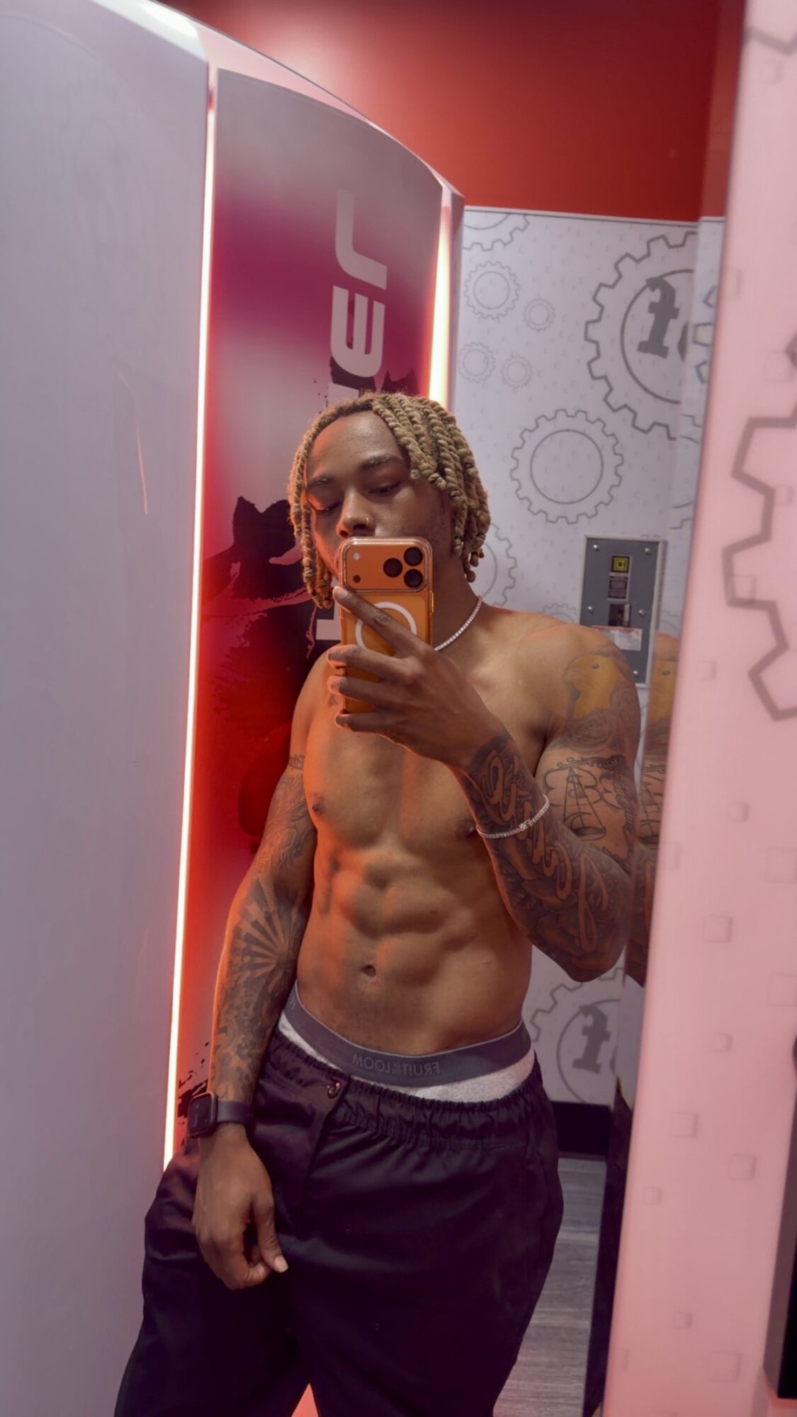 Young man with curly hair taking a mirror selfie, shirtless, showing muscular abs, in a room with red and pink walls.