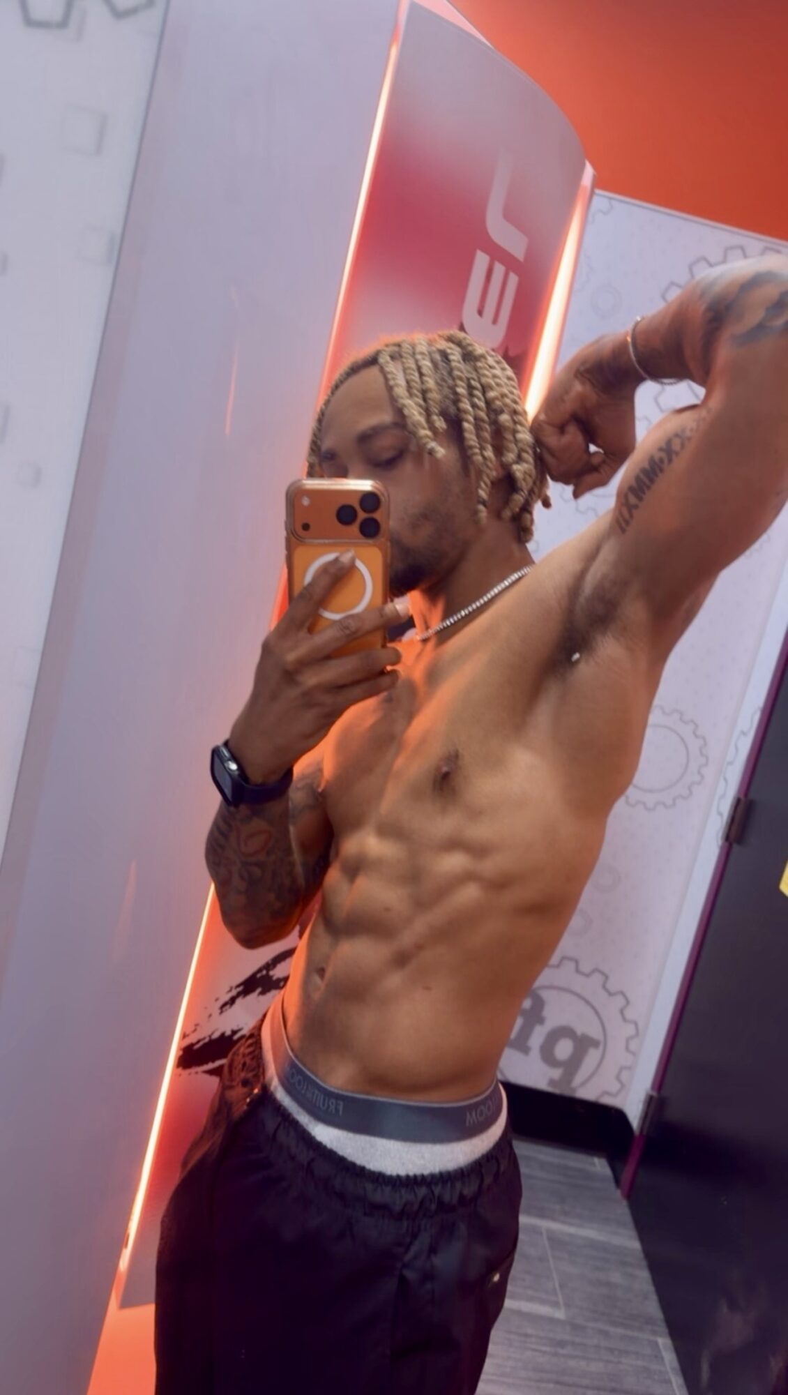 Young man with braided hair taking a mirror selfie, shirtless, flexing muscles, in a gym or fitness center.