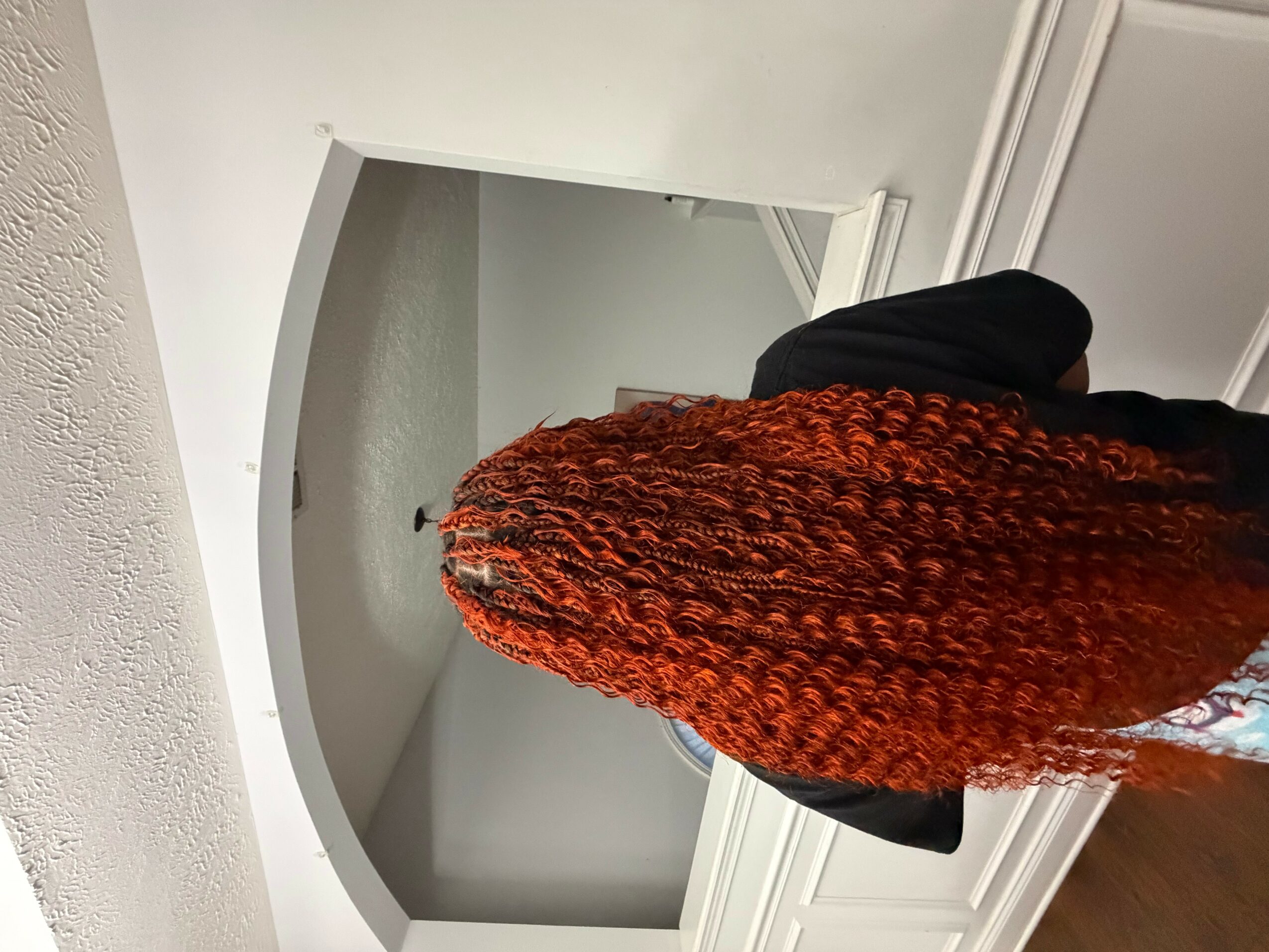 Person with long, curly, reddish-brown hair standing indoors, facing away from the camera, in front of a mirror.