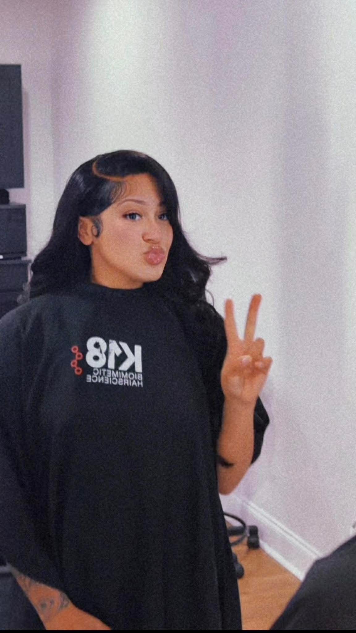 Young woman with dark hair making a peace sign gesture, wearing a black shirt, standing indoors against a white wall.