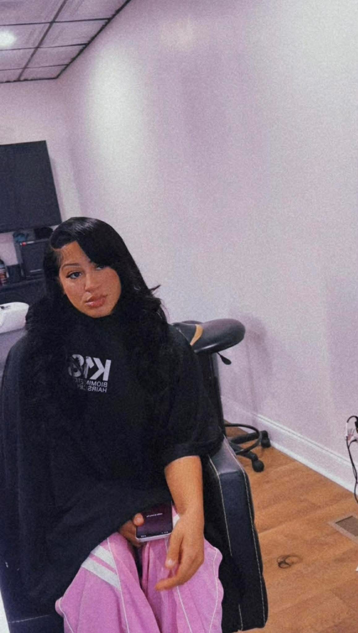 Young woman with black hair sitting in a chair, holding a phone, in a room with white walls and wooden floor.