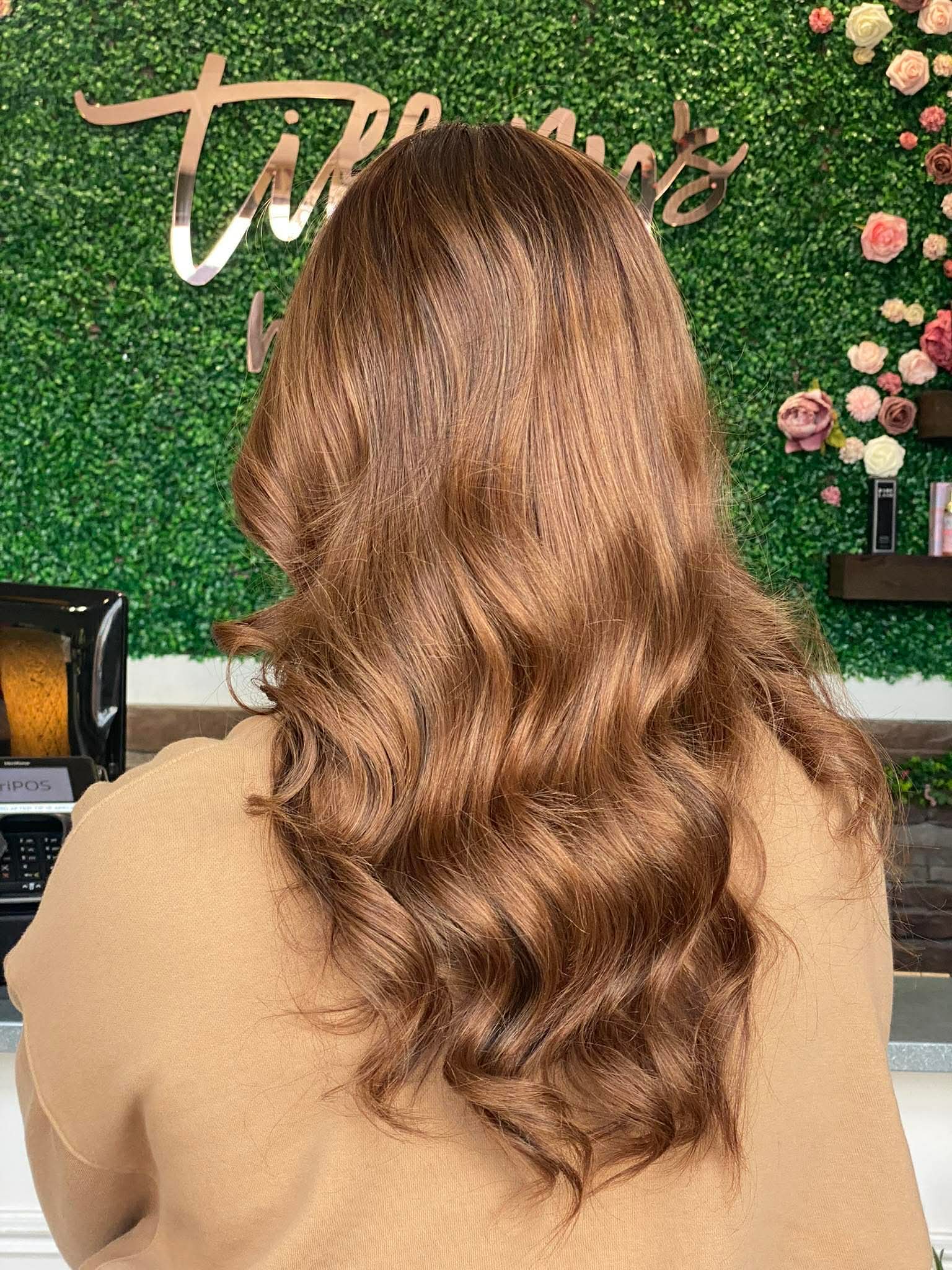 Back of a woman with long, wavy, light brown hair, standing in front of a green floral wall with pink flowers.
