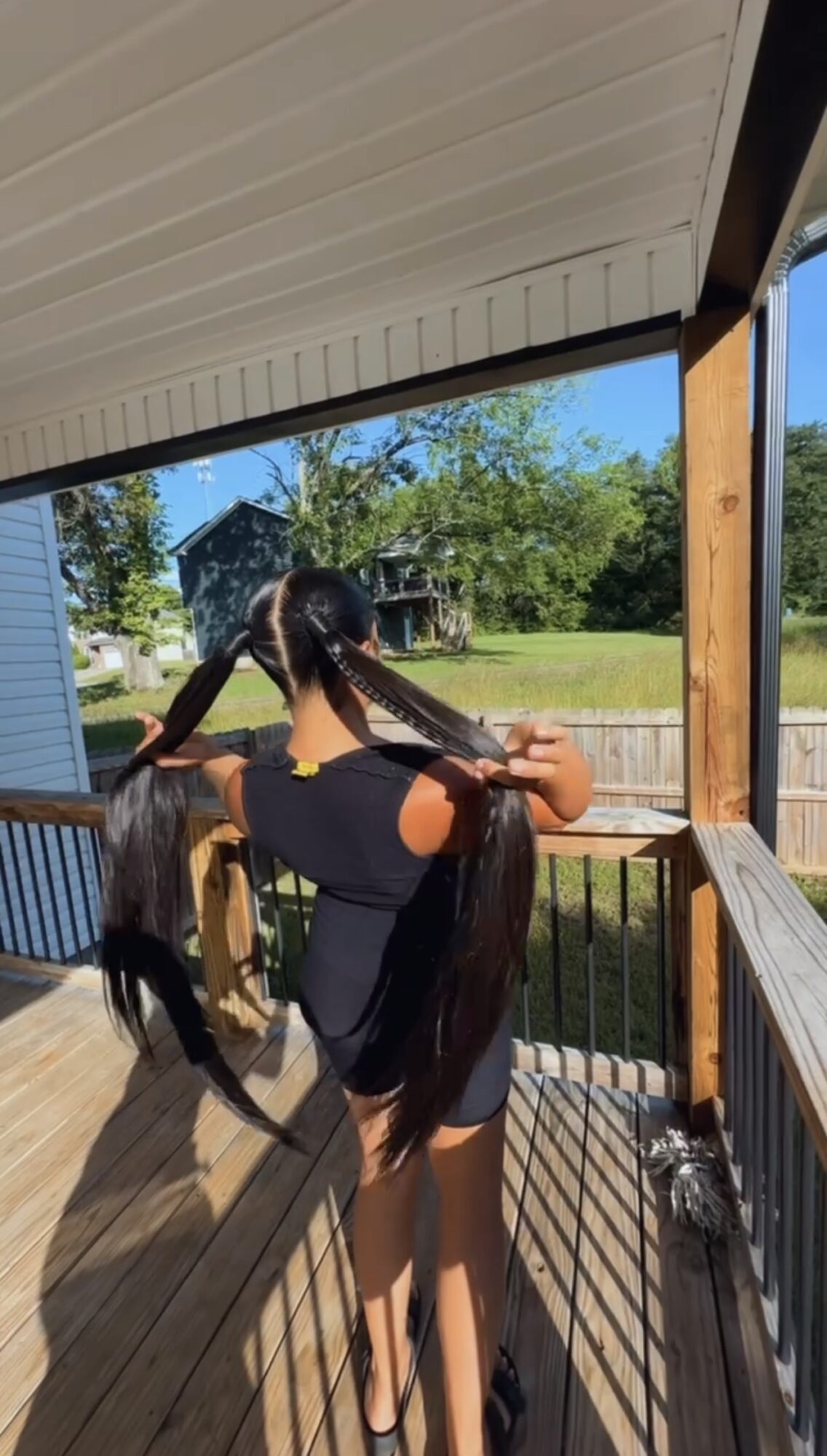 Person with long hair standing on porch, holding hair, overlooking yard with trees and houses, sunny day.
