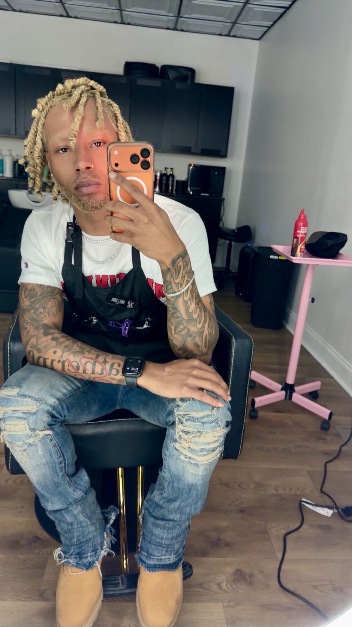 Person with blonde dreadlocks taking a mirror selfie in a salon, wearing a white shirt, ripped jeans, and tan shoes.