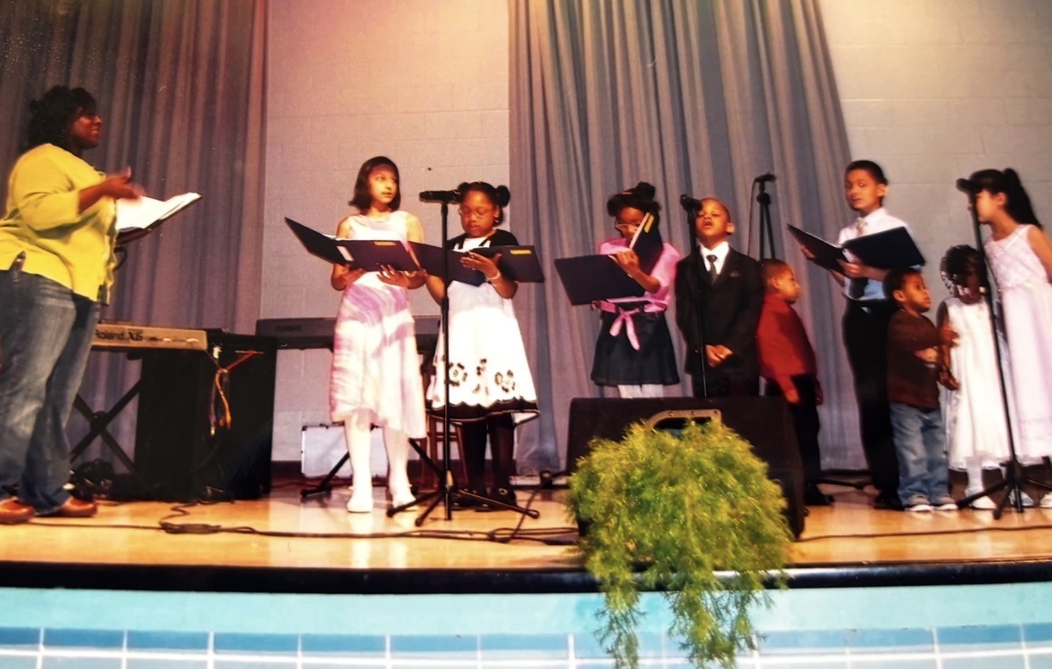 Children and a woman on stage holding music sheets, with a curtain backdrop and a potted plant in front.
