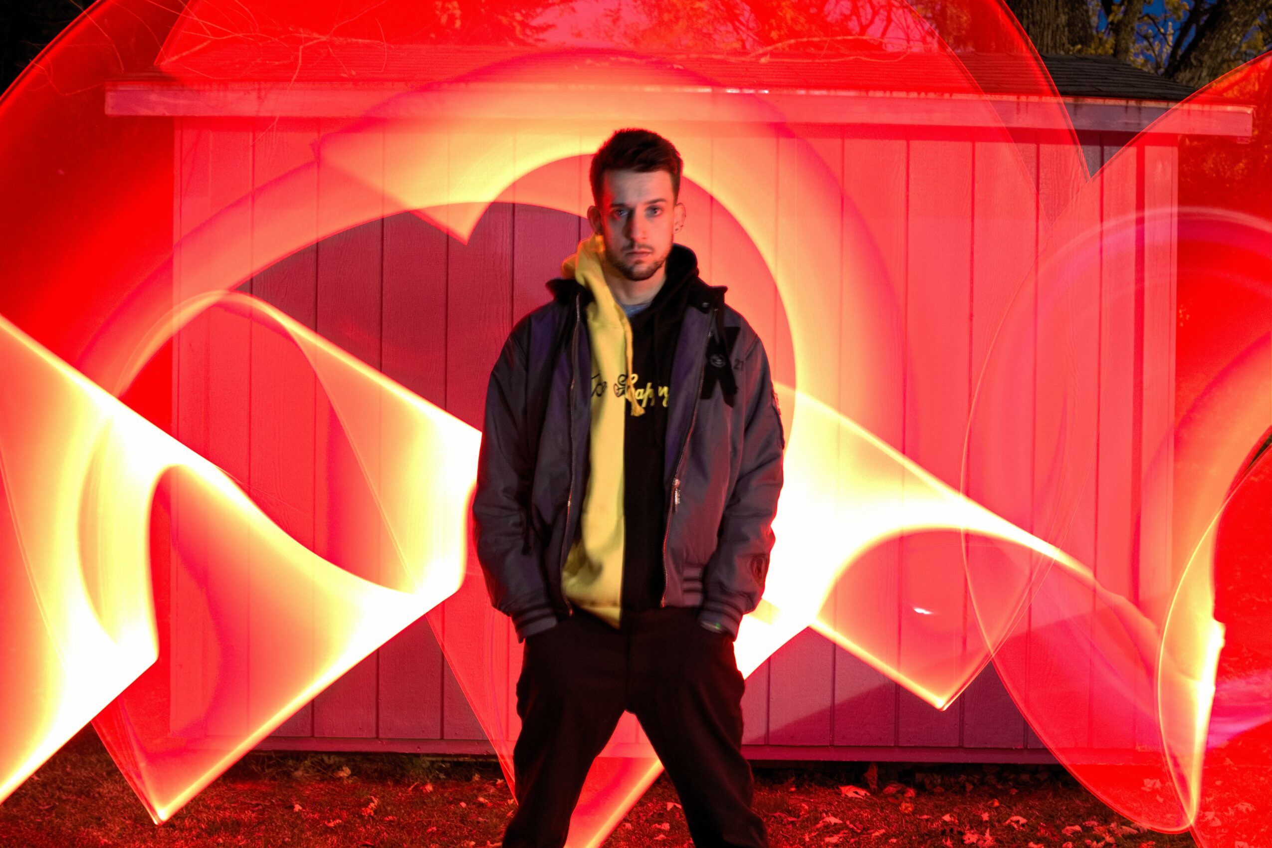Young man standing in front of a red fence with abstract red and yellow light patterns behind him.
