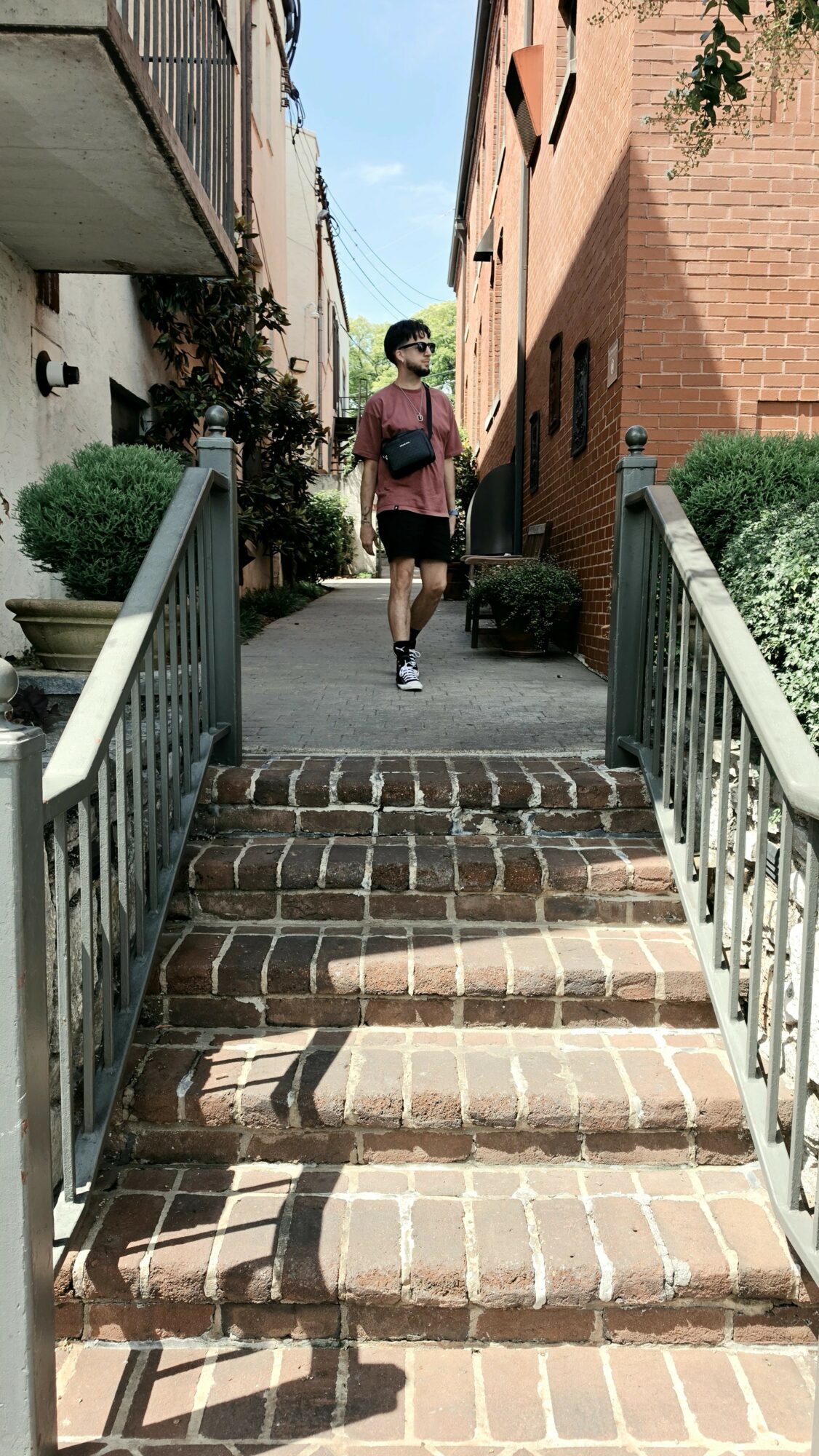 Person walking up brick stairs between buildings, with railings on both sides, greenery, and a blue sky.