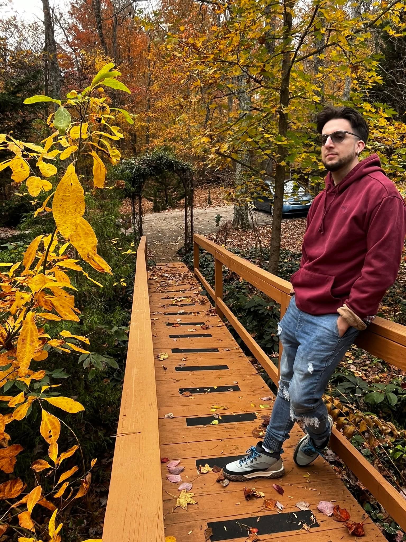 Man in maroon hoodie and jeans standing on a wooden bridge in a park with autumn trees and fallen leaves.