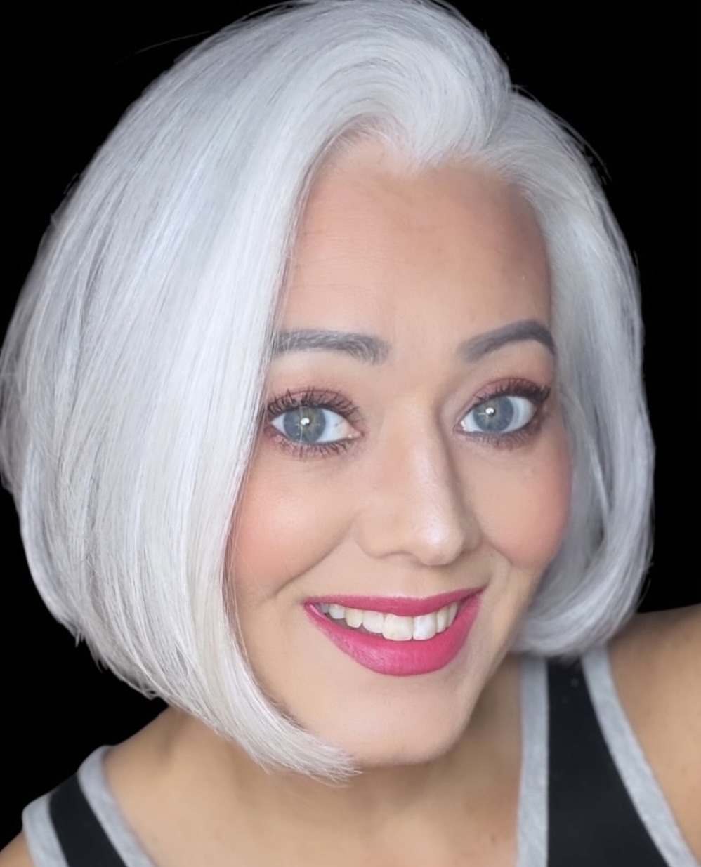 Portrait of a woman with short, white hair, blue eyes, and pink lipstick, smiling against a black background.