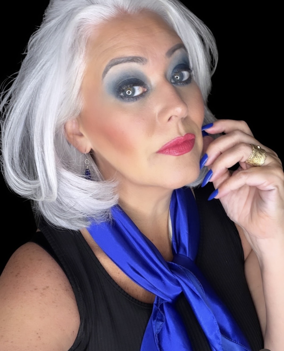 Woman with silver hair, bold makeup, blue eyeshadow, and a blue scarf, looking at the camera with a hand near her face.