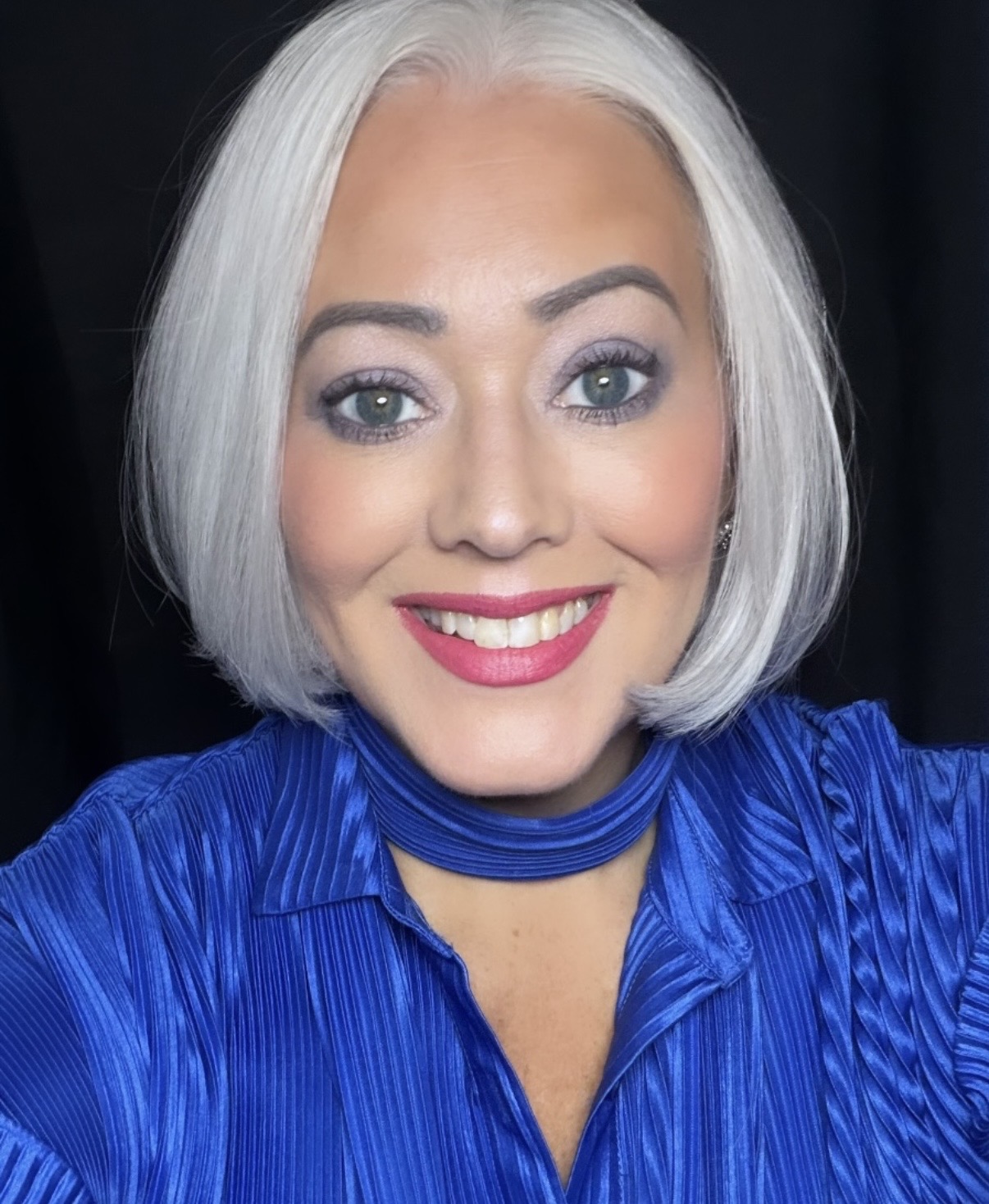Woman with short white hair, blue eyes, wearing a blue top, smiling, against a dark background.