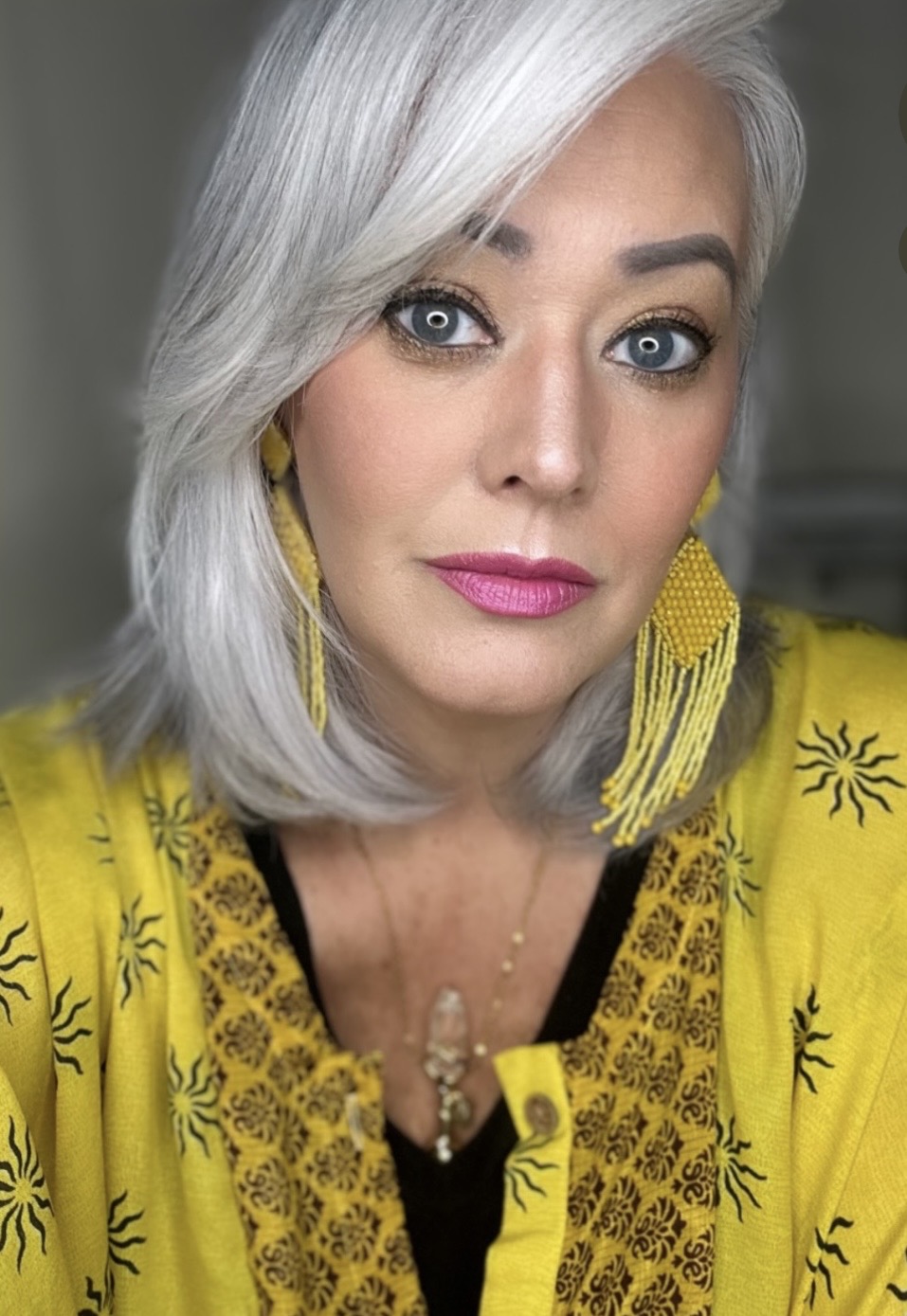 Portrait of a woman with silver hair, blue eyes, wearing yellow earrings and a yellow top with black embroidery.