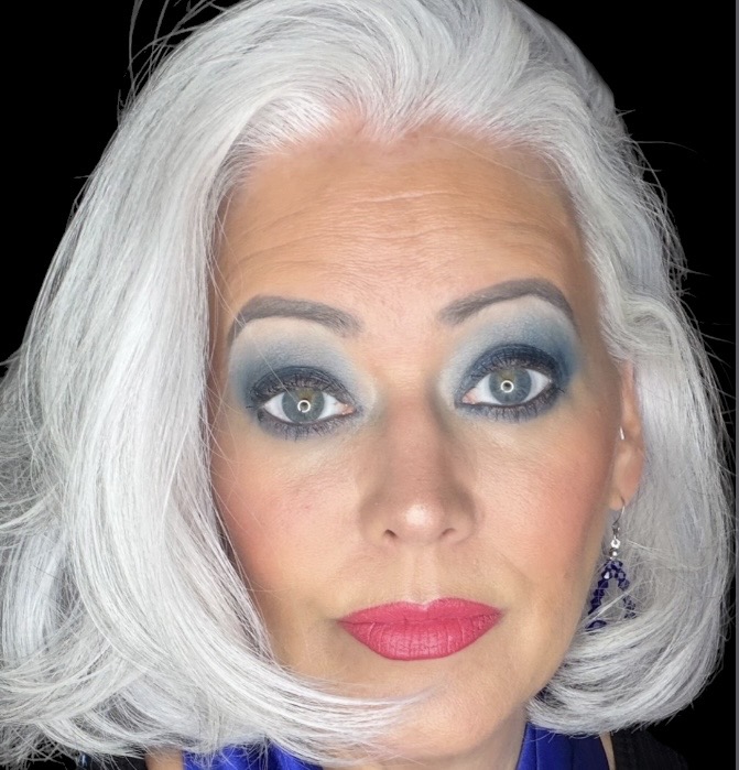 Portrait of a woman with white hair, bold eye makeup, and pink lipstick, looking directly at the camera.