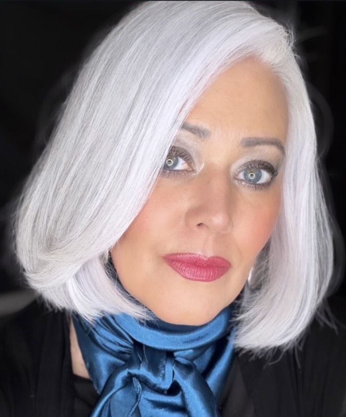 Woman with white hair, blue eyes, wearing a blue scarf and black top, looking at the camera.