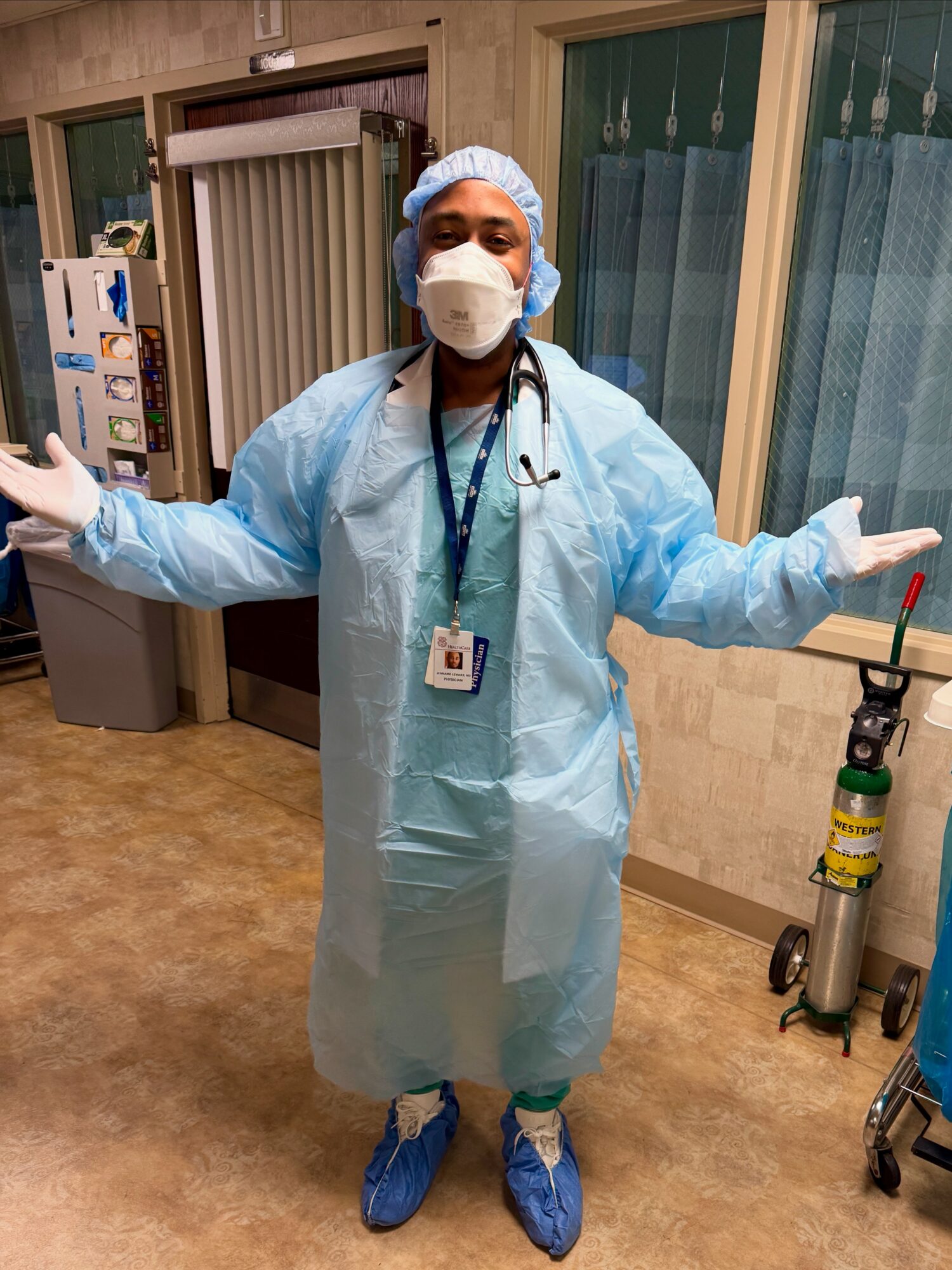 Healthcare worker in protective gear with arms outstretched in a hospital room.