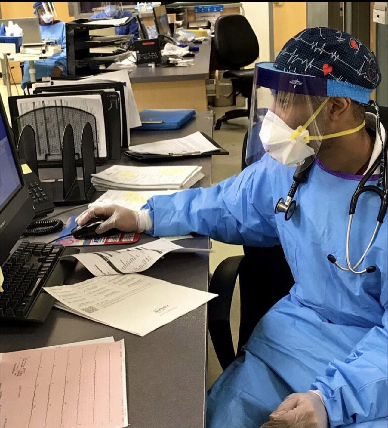 Healthcare worker in blue gown, face shield, mask, and gloves working at a desk with papers and a computer.