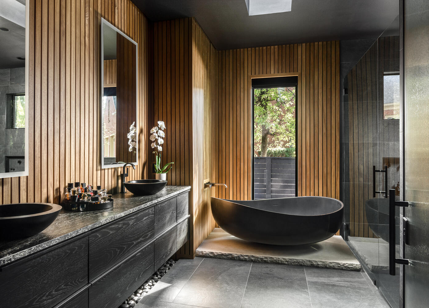 Modern bathroom with black bathtub, wooden wall panels, and a large window showing outdoor greenery.