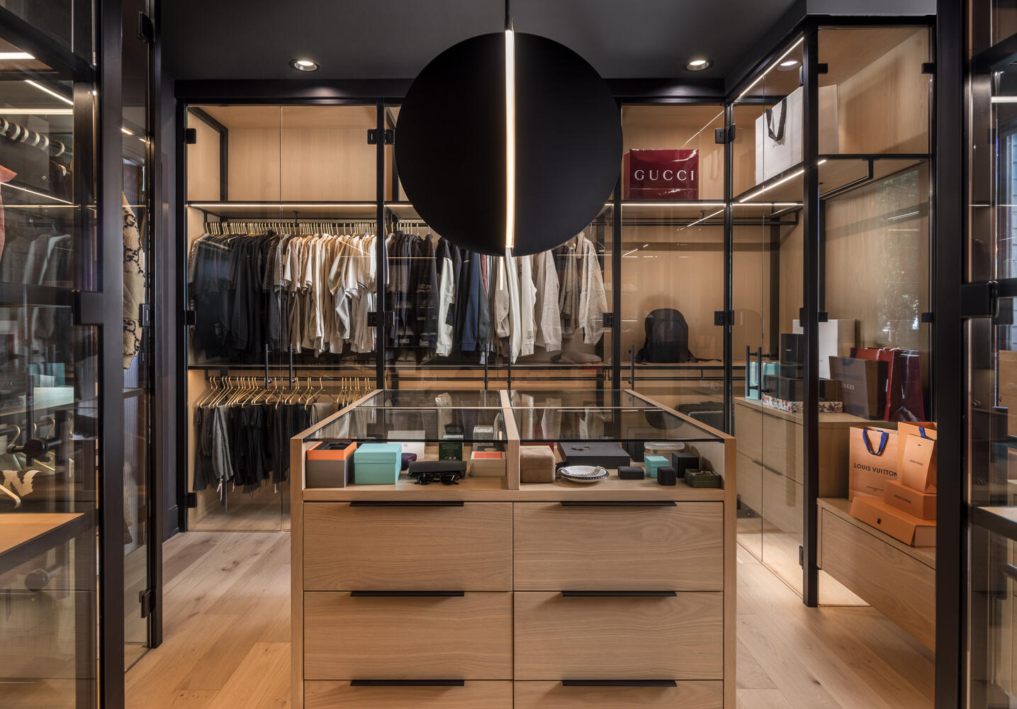 Closet with clothing racks, glass display case, and shelves with designer bags and accessories, illuminated by spotlights.