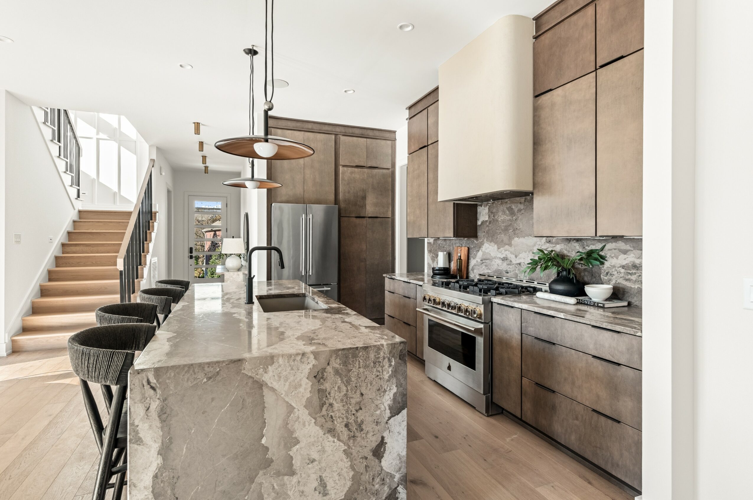 Modern kitchen with island, wooden cabinets, stainless steel appliances, and a staircase in the background.