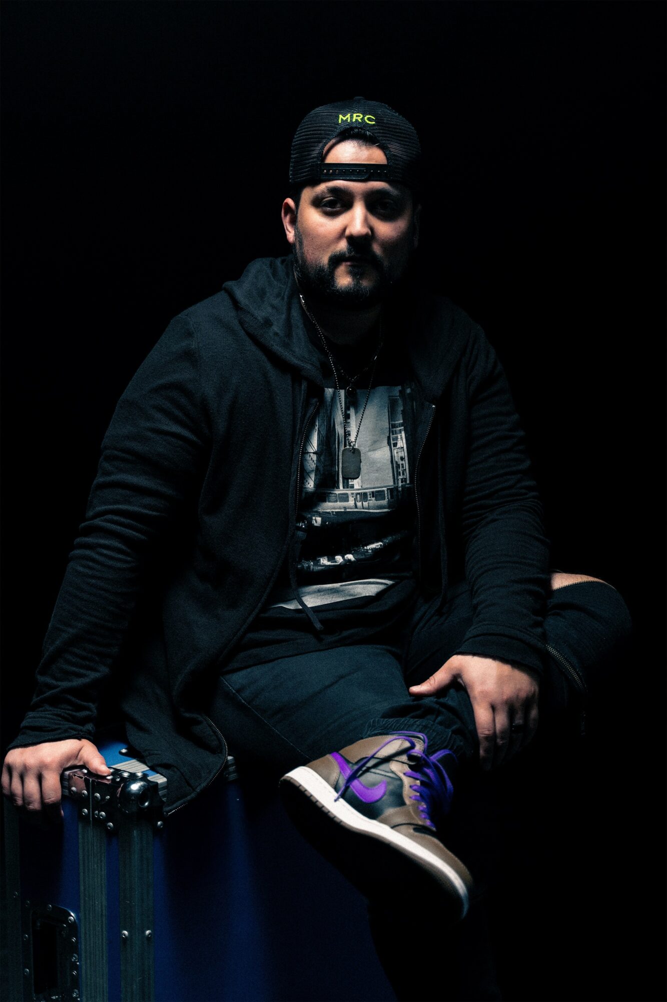 Person sitting in a dark setting, wearing a black cap, black jacket, and sneakers, with a serious expression.