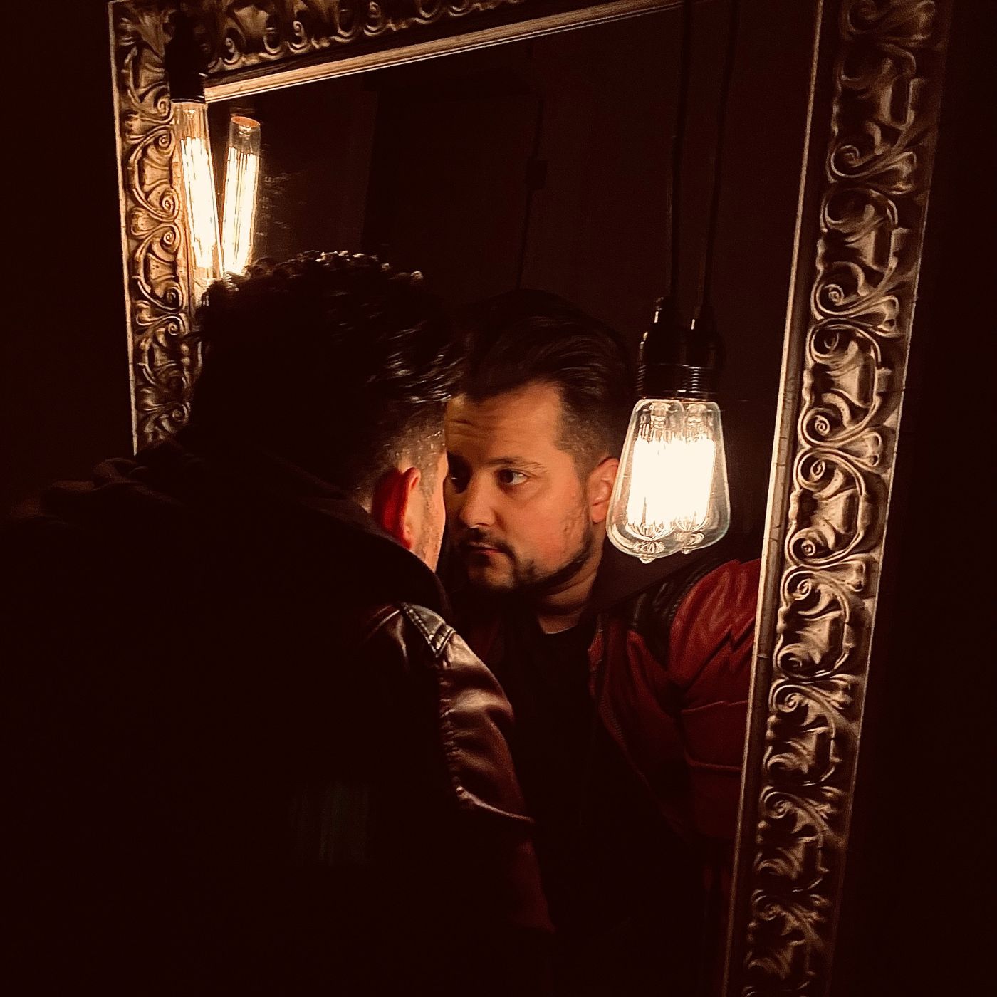 Two men looking at each other in a mirror with ornate frame, warm lighting, and dark background.
