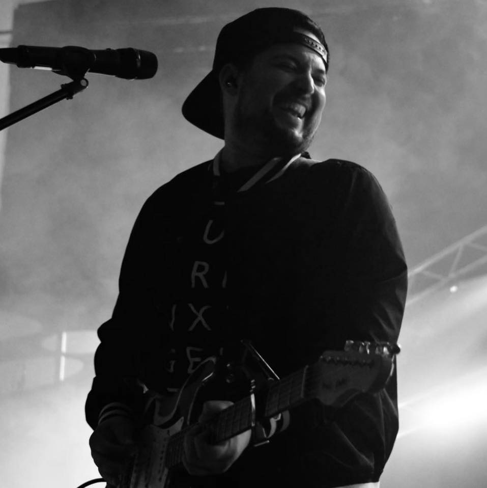 Person playing guitar on stage, smiling, wearing a cap backwards, with microphone nearby, in black and white.