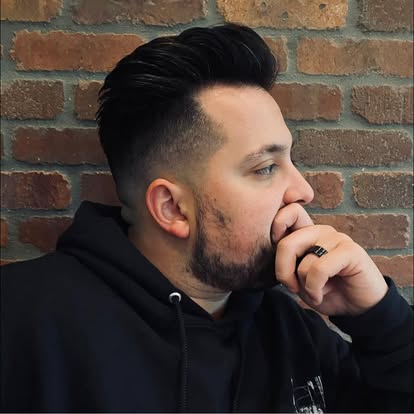 Side profile of a man with dark hair and beard, wearing a black hoodie, against a brick wall.