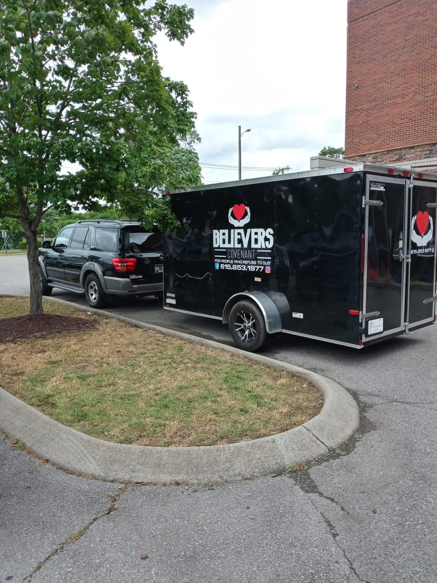 Black trailer with 'BELIEVERS' logo parked next to a black vehicle, near a tree and a brick building.