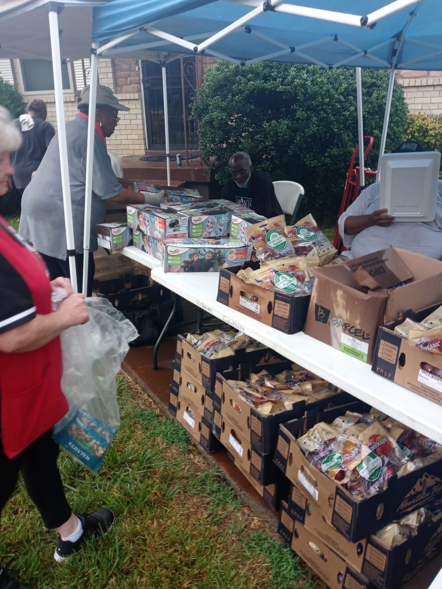 People gather around a table with boxes of food under a blue canopy outdoors, with a brick building and greenery in background.