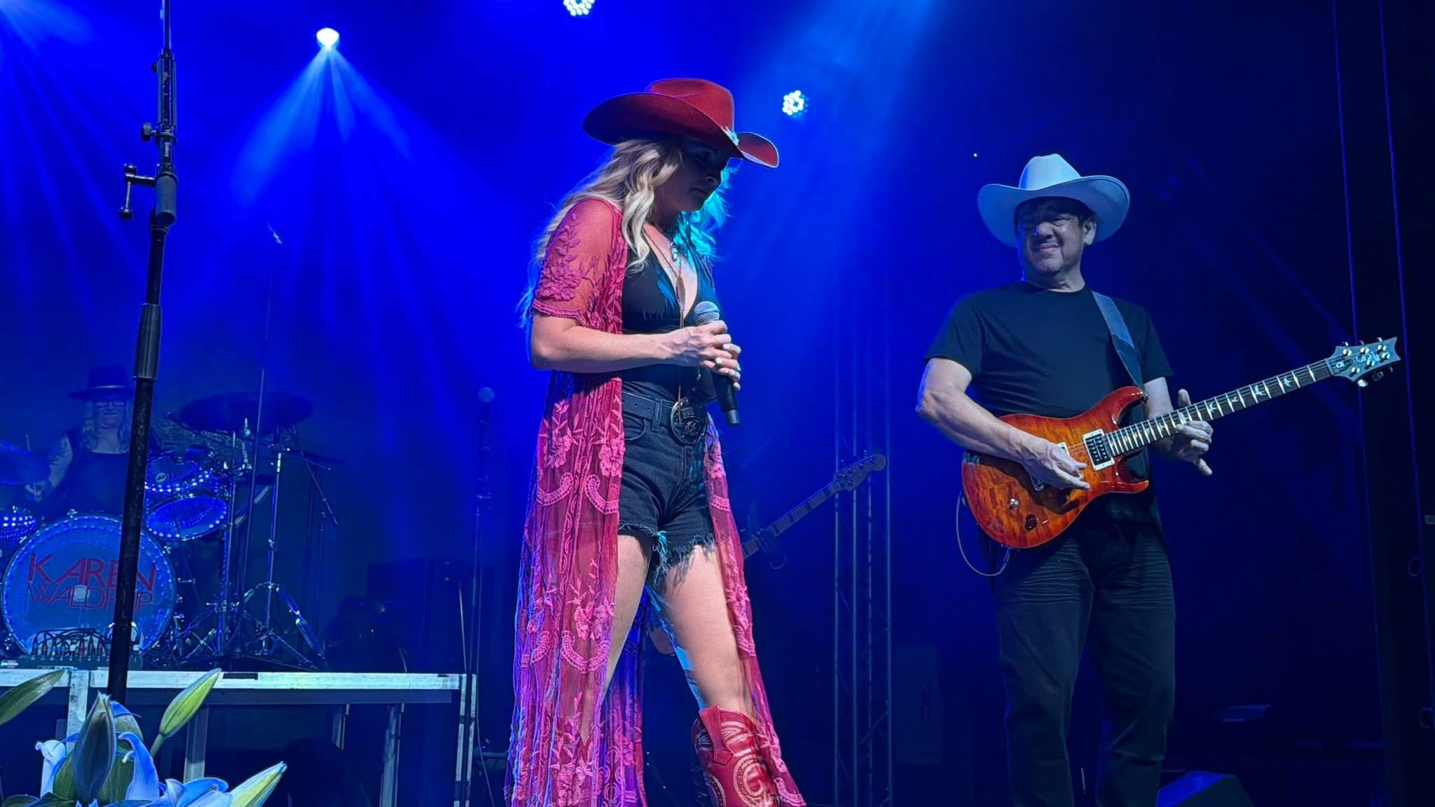 Two musicians perform on stage with blue lighting, one singing and the other playing guitar, in a concert setting.