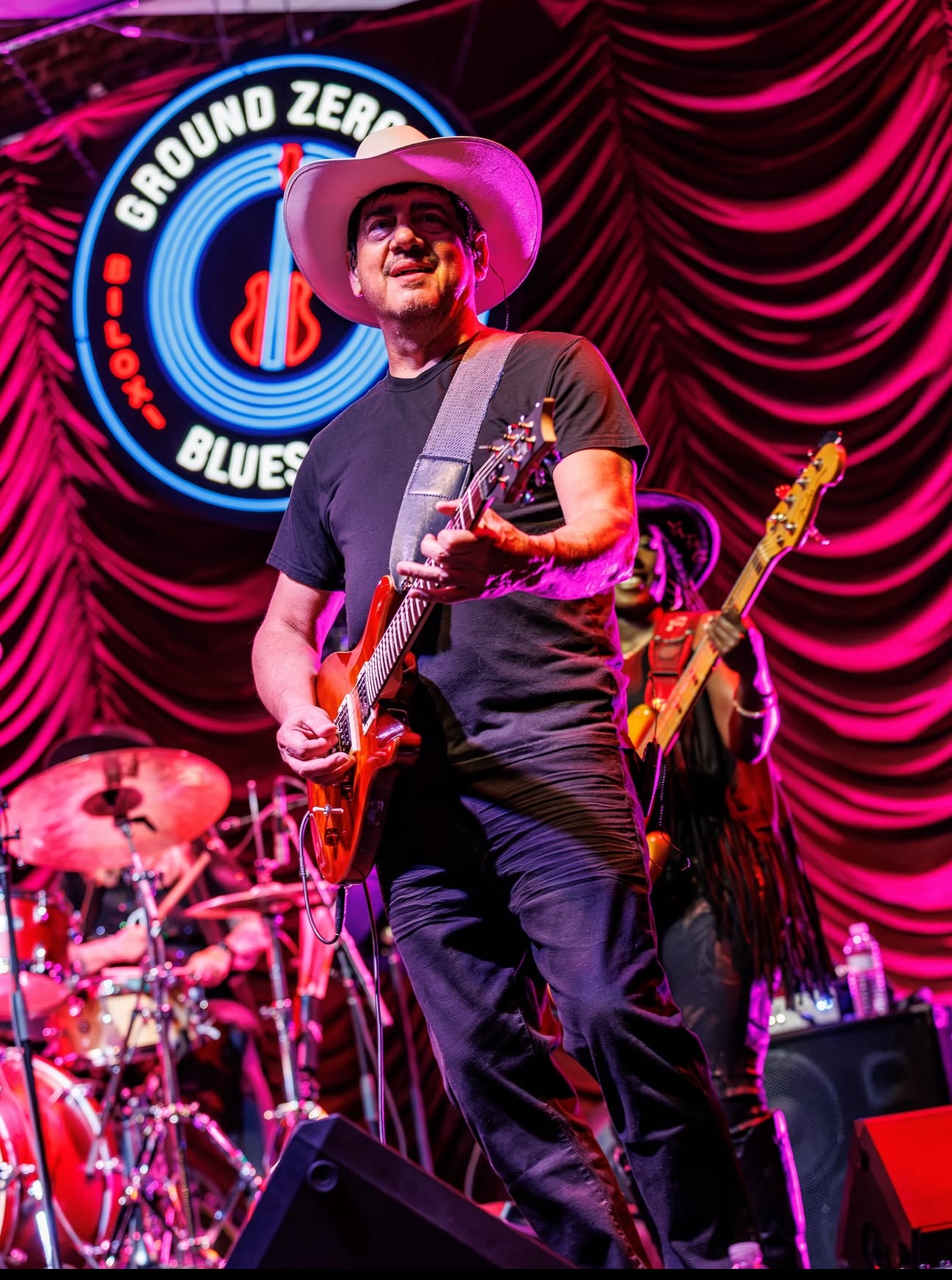 Musician wearing a large hat playing guitar on stage with red curtains and a drum set in the background.